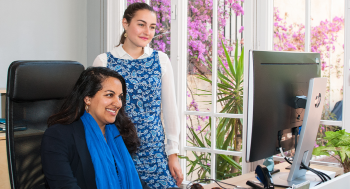 A woman sitting in front of a desk with a computer next to another woman who is standing up